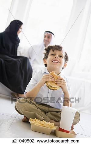 Stock Image - Young boy with fast food in living room laughing with parents in background (high key/selective focus). Fotosearch