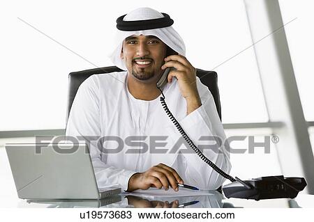 Stock Image - Businessman in office with laptop on telephone smiling (high key/selective focus). Fotosearch