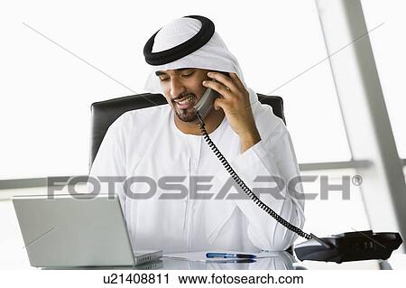 Stock Image - Businessman in office with laptop on telephone smiling (high key/selective focus). Fotosearch