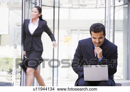 Picture - Businessman outdoors in front of building using laptop with businesswoman in background smiling (high key/blur). Fotosearch