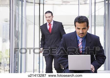 Stock Photo - Businessman outdoors in front of building using laptop with businessman in background (high key/selective focus). Fotosearch