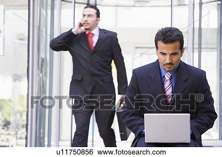 Stock Photograph - Businessman outdoors in front of building using laptop with businessman in background on cellular phone (high key/selective focus). Fotosearch