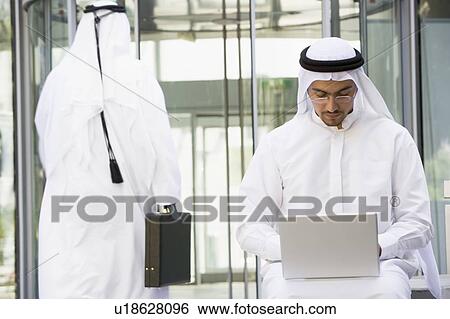 Stock Photograph - Businessman outdoors using laptop with businessman walking in background (selective focus). Fotosearch