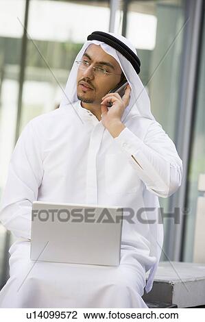 Stock Image - Businessman outdoors with laptop using cellular phone by building (selective focus). Fotosearch