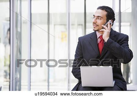 Picture - Businessman sitting outdoors by building with a laptop on cellular phone (high key/selective focus). Fotosearch