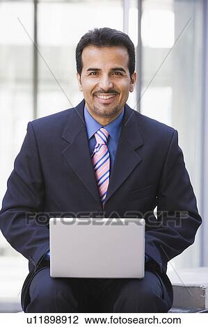 Stock Image - Businessman sitting outdoors by building with a laptop smiling (high key/selective focus). Fotosearch