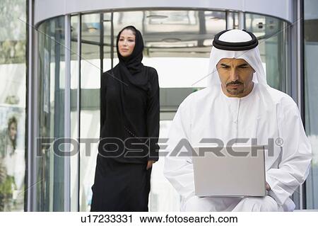 Stock Image - Businessman sitting outdoors by building using laptop with businesswoman walking in background (selective focus). Fotosearch