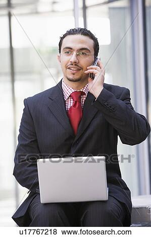 Stock Photo - Businessman sitting outdoors by building with a laptop on cellular phone (high key/selective focus). Fotosearch