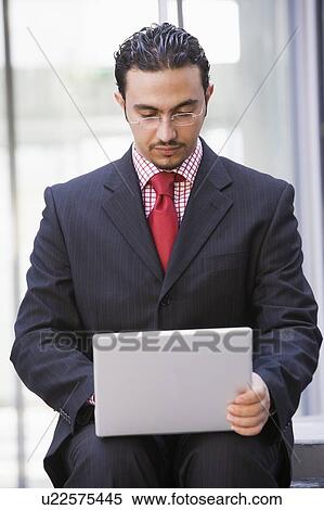 Stock Photography - Businessman sitting outdoors by building with a laptop (high key/selective focus). Fotosearch