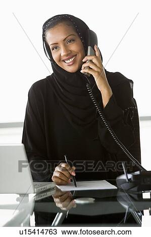 Stock Image - Businesswoman in office by laptop on telephone smiling (high key/selective focus). Fotosearch