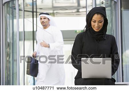 Stock Image - Businesswoman sitting outdoors by building with laptop smiling with businessman running in background (selective focus). Fotosearch