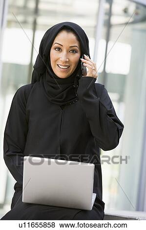Stock Photo - Businesswoman sitting outdoors by building with laptop using cellular phone smiling (selective focus). Fotosearch