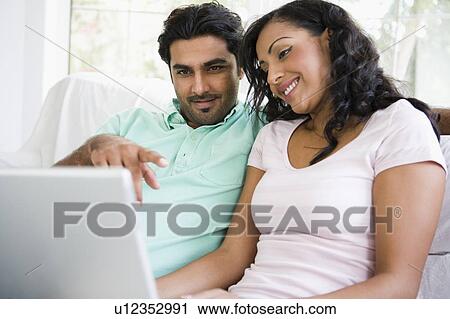Stock Image - Couple in living room with laptop pointing and smiling (high key/selective focus). Fotosearch