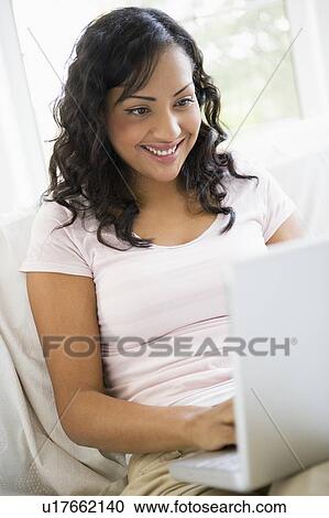 Stock Image - Woman in living room with laptop smiling (high key/selective focus). Fotosearch