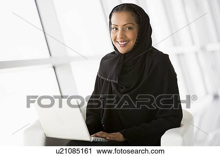 Stock Image - Woman sitting indoors with laptop smiling (high key/selective focus). Fotosearch