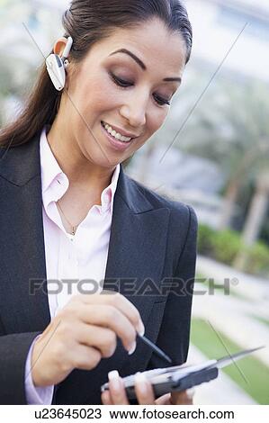 Stock Image - Woman wearing headset outdoors and using personal digital assistant smiling (selective focus). Fotosearch