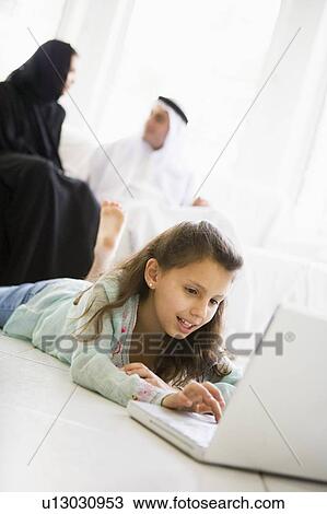 Stock Image - Young girl in living room using laptop and smiling with parents in background (high key/selective focus). Fotosearch