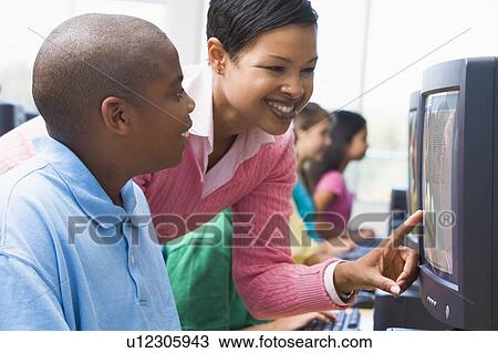 Stock Image - Teacher helping student at computer terminal with students in background (depth of field/high key). Fotosearch