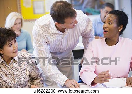 Stock Image - Adult students in class with teacher helping one (selective focus). Fotosearch