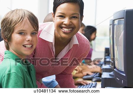 Picture - Teacher helping student at computer terminal with students in background (depth of field/high key). Fotosearch