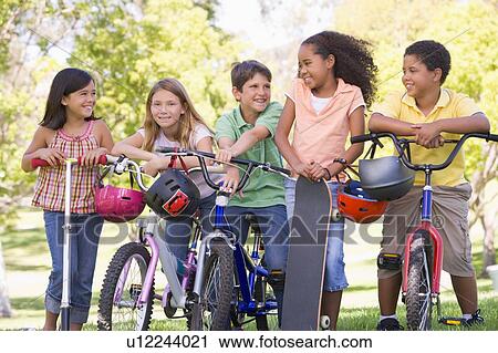 Stock Image - Five young friends with bicycles scooters and skateboard outdoors smiling. Fotosearch