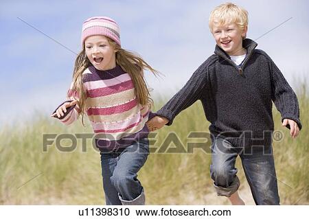 Stock Image - Two young children running on beach holding hands smiling. Fotosearch