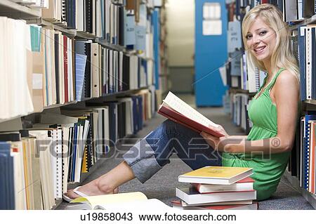 Stock Image - Woman sitting on floor in library holding book. Fotosearch