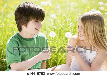 Stock Image - Two young children sitting outdoors holding dandelion heads smiling. Fotosearch