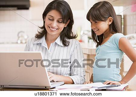 Stock Image - Woman and young girl in kitchen with laptop and paperwork smiling. Fotosearch