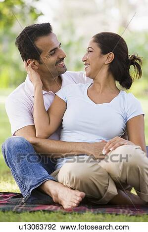Stock Image - Couple sitting outdoors in park being affectionate and smiling (selective focus). Fotosearch