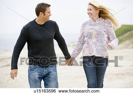 Stock Photograph - Couple walking at beach holding hands smiling. Fotosearch
