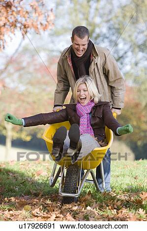 Stock Image - Man outdoors pushing woman in wheelbarrow and smiling (selective focus). Fotosearch
