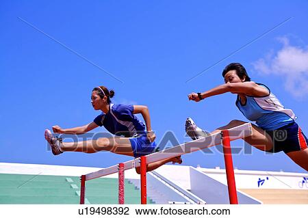 Stock Image - Two female athletes jumping hurdles. Fotosearch