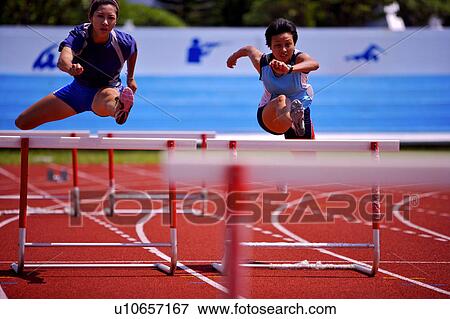 Stock Photo - Two female athletes jumping hurdles, front view. Fotosearch