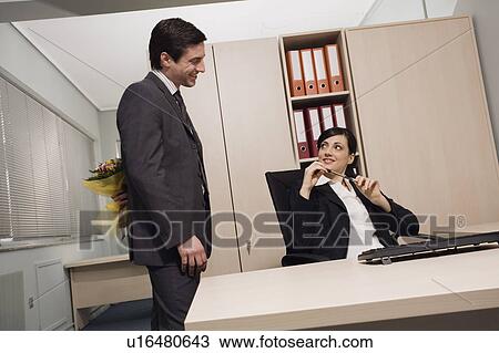 Stock Image - Male office worker hiding flowers for female colleague. Fotosearch