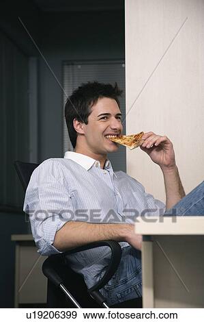 Stock Photo - Office worker eating a pizza slice at desk. Fotosearch