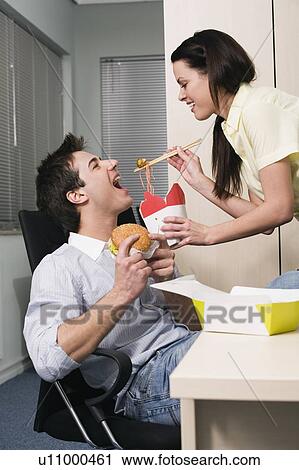 Stock Image - Office worker feeding her colleague at desk. Fotosearch