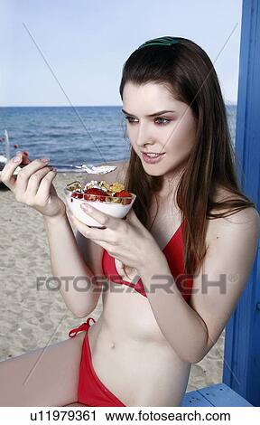 Stock Image - Female young adult eating fruit and yogurt on beach. Fotosearch