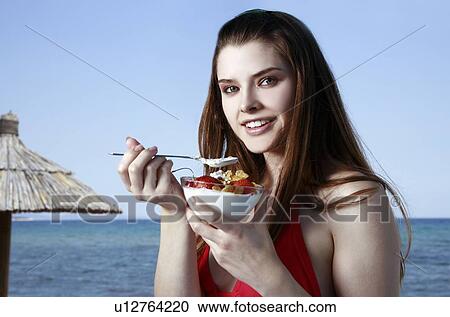 Stock Image - Female young adult eating fruit and yogurt on beach. Fotosearch