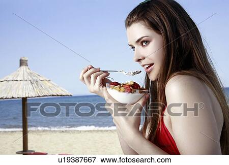 Stock Photo - Female young adult eating fruit and yogurt on beach. Fotosearch