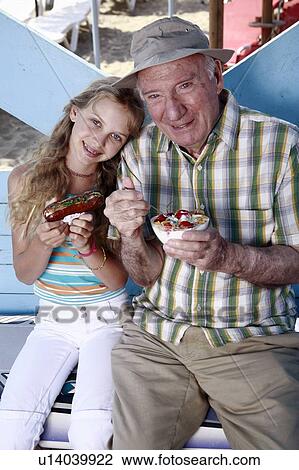 Stock Image - Grandfather and granddaughter eating at beach. Fotosearch