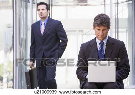 Businessman outdoors in front of building using laptop with businessman in background (high key/selective focus) View Large Photo Image Stock Photo - Businessman outdoors in front of building using laptop with businessman in background (high key/selective focus). Fotosearch