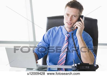Businessman sitting in office with laptop using telephone View Large Photo Image Stock Photo - Businessman sitting in office with laptop using telephone. Fotosearch
