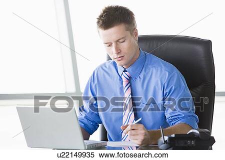 Stock Photography - Businessman sitting in office with laptop writing note. Fotosearch