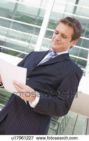 Businessman standing outdoors looking at paperwork View Large Photo Image Stock Photo - Businessman standing outdoors looking at paperwork. Fotosearch