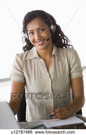 Businesswoman wearing headset in office by laptop smiling (high key/selective focus) View Large Photo Image Stock Photograph - Businesswoman wearing headset in office by laptop smiling (high key/selective focus). Fotosearch