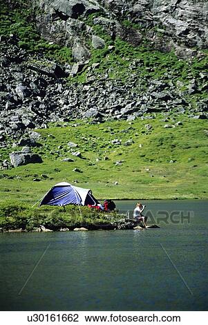 Stock Image - Camper outdoors at campsite by lake drinking beverage (far away). Fotosearch