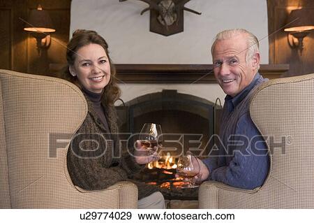 Stock Photo - Couple sitting in living room by fireplace with drinks smiling. Fotosearch