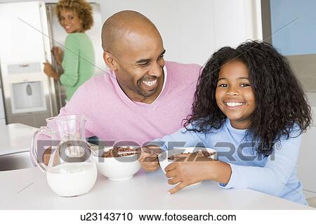 Stock Image - Father Sitting With Daughter As She They Eat Breakfast With Her Mother In The Background . Fotosearch
