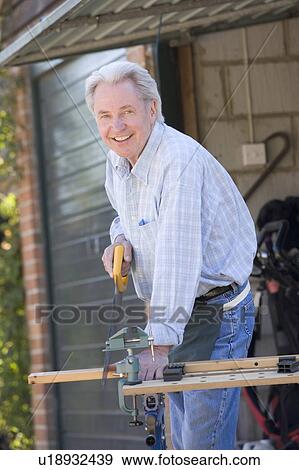 Stock Photo - Man at shed sawing wood and smiling. Fotosearch